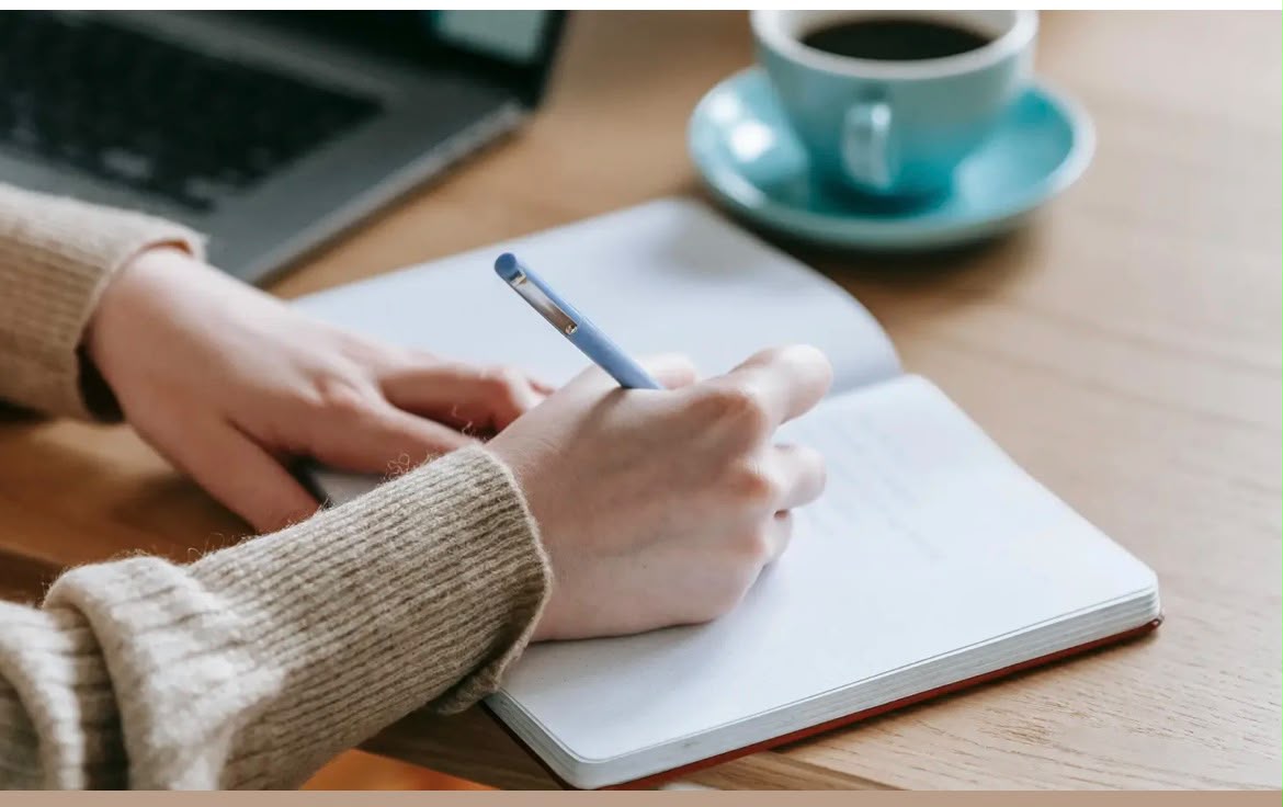A person writing notes in a notebook beside a laptop